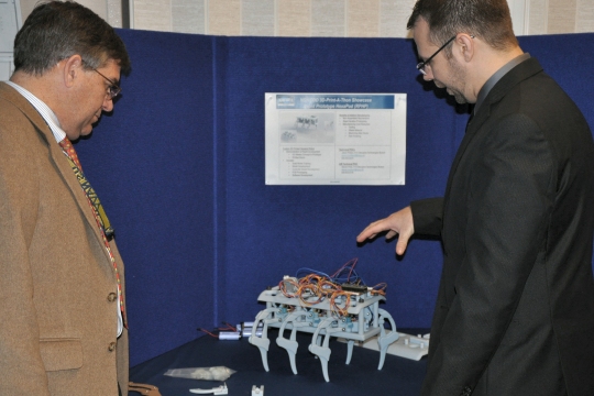 Jason Phillips (right), a Naval Surface Warfare Center Dahlgren Division (NSWCDD) engineer, demonstrates the Rapid Prototype HexaPod Robot for NSWCDD Technical Director Dennis McLaughlin. It was showcased at the command's Print-A-Thon in December 2015. The reality of 3D Printing - also known as additive manufacturing - is expanding across the Navy's science and engineering community via Naval Surface Warfare Center Dahlgren Division (NSWCDD) and Combat Direction Systems Activity (CDSA) Dam Neck. (U.S. Navy photo by Luefras Robinson/Released)