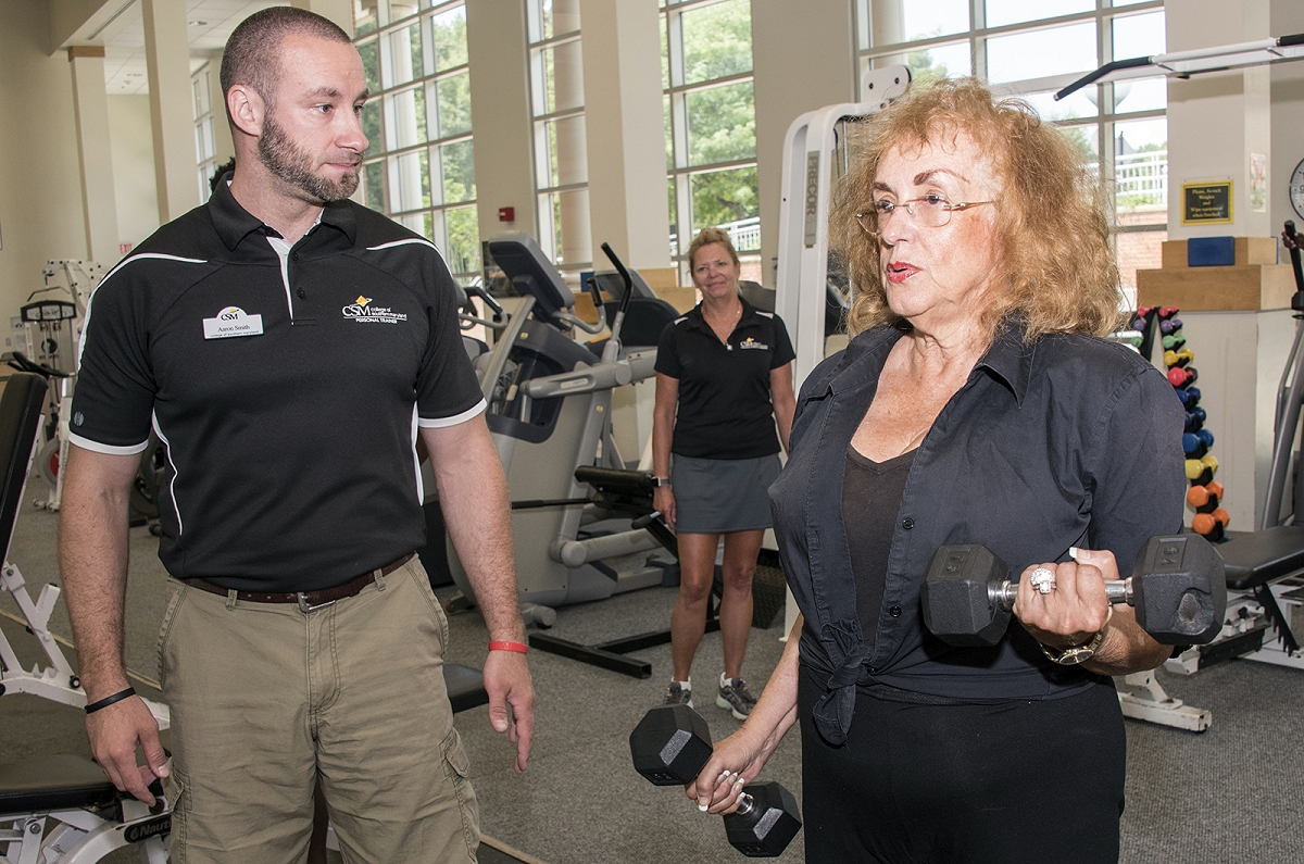 CSM Fitness Trainer Aaron Smith, left, checks Charlotte Weirich on her lifting technique at the La Plata Campus fitness center, as CSM Fitness Coordinator Jane Pomponio observes from behind.